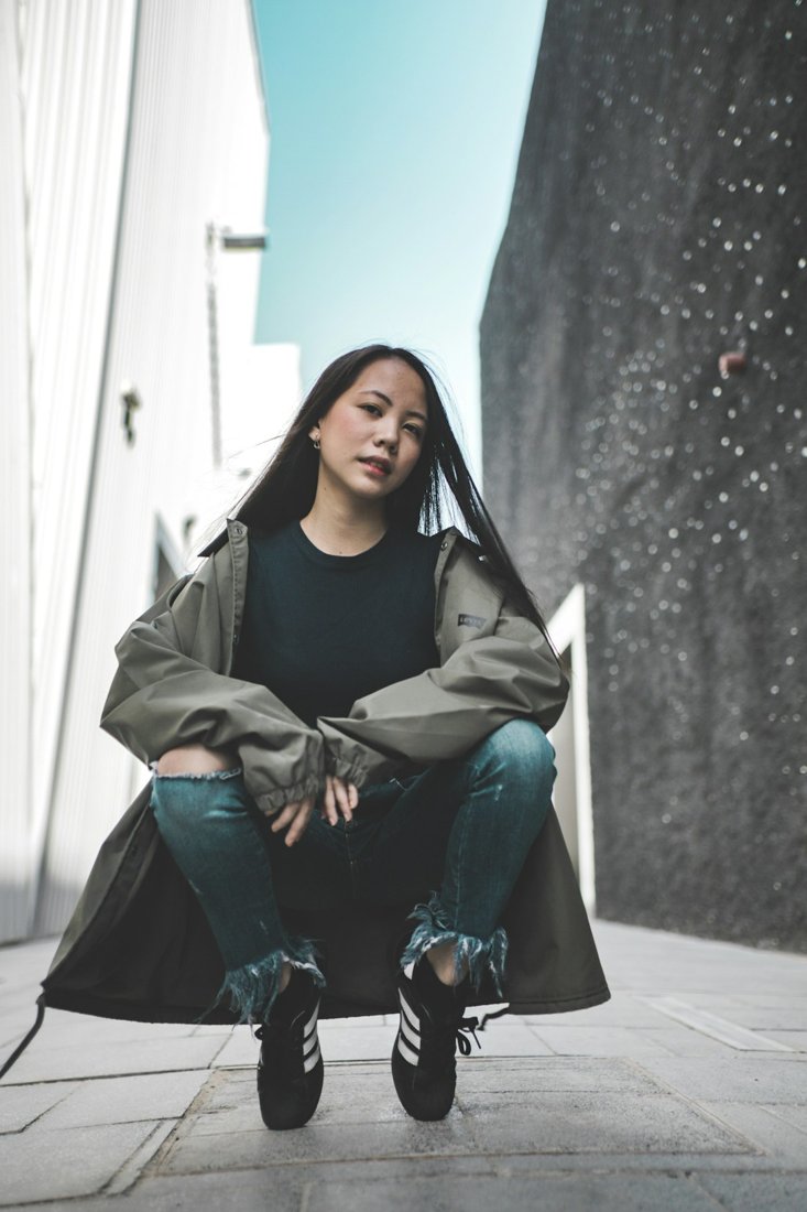 A woman crouching on a city pavement in a green coat, jeans and black ankle boots