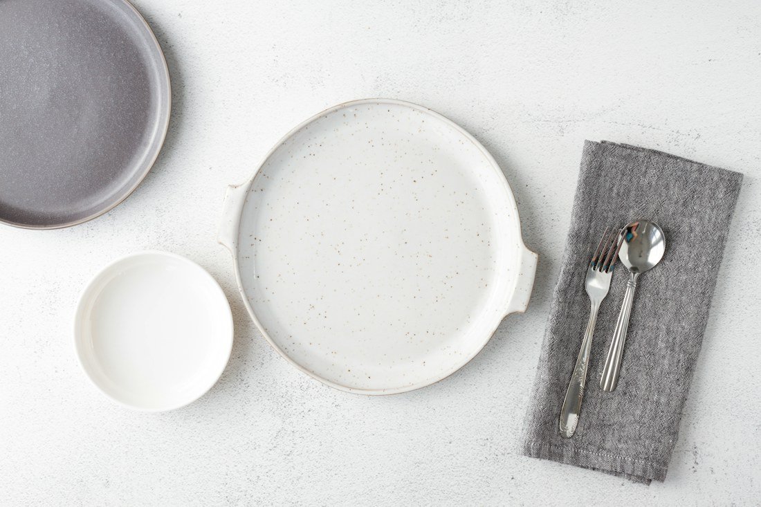 White and gray ceramic plates beside a fork and spoon on a folded gray linen napkin