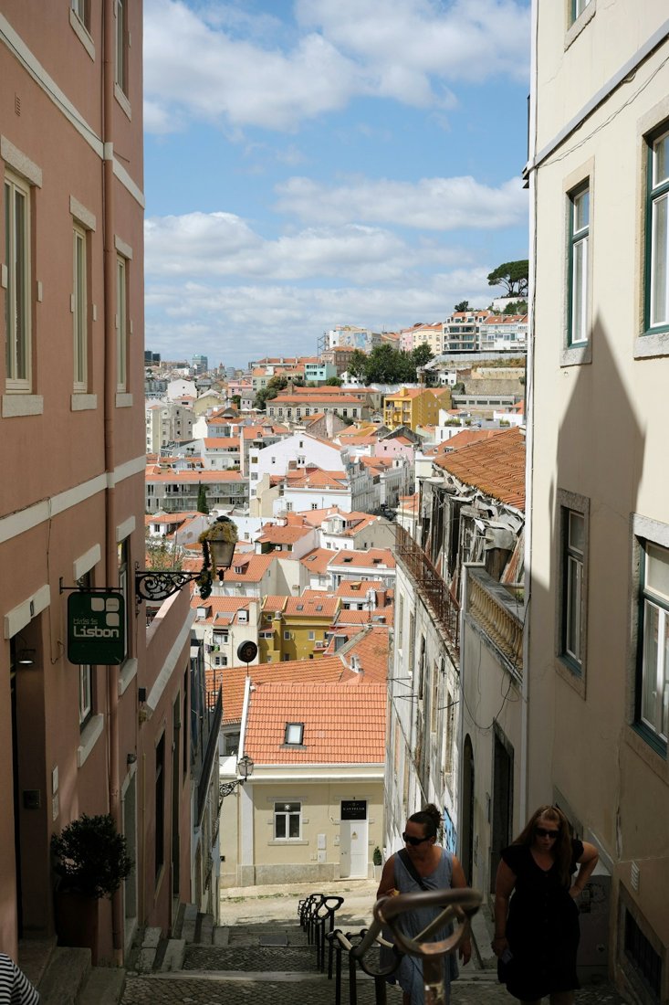 People on a steep Lisbon stairway with red rooftops visible beyond