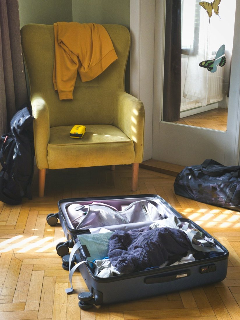 An open packed suitcase on a sunlit wooden floor beside a yellow armchair